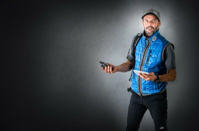 Wasti Obermoser Ein Mann in einer blauen Weste steht vor einem grauen Hintergrund. Er hält ein mobiles Gerät in einer Hand und ein Tablet in der anderen.