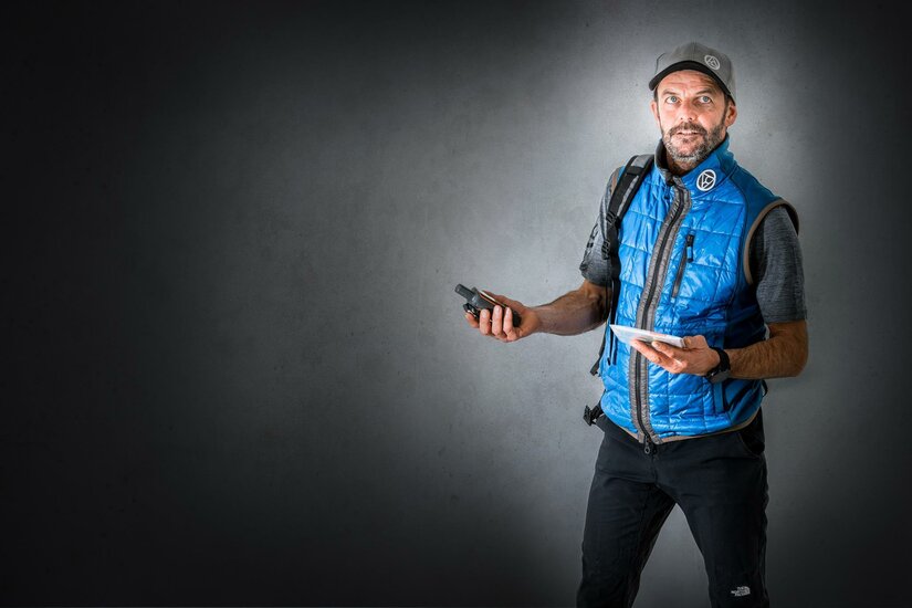 Wasti Obermoser Ein Mann in einer blauen Weste steht vor einem grauen Hintergrund. Er hält ein mobiles Gerät in einer Hand und ein Tablet in der anderen.