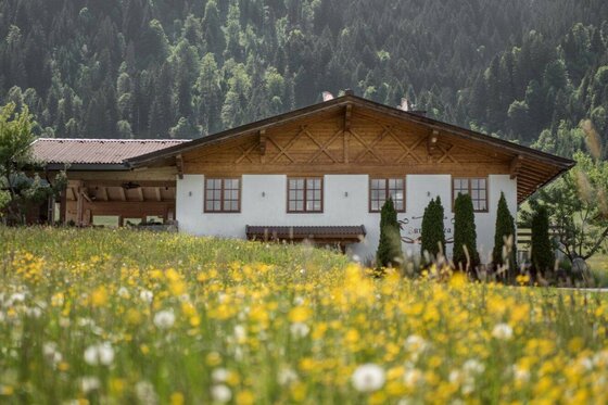 Ein weißes Gebäude mit Holzgiebel hinter einer blühenden Blumenwiese vor bewaldetem Hintergrund.