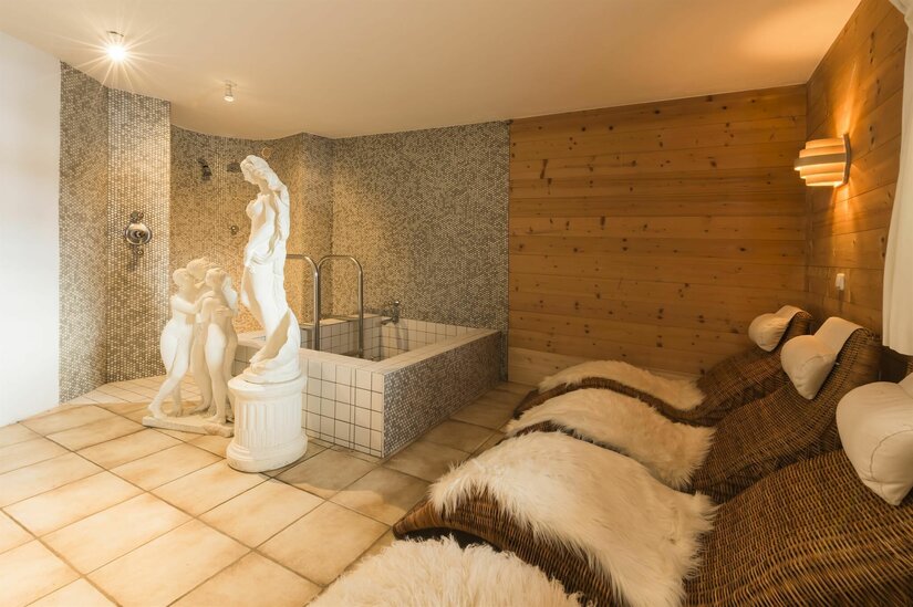 Wellnessbereich Das Kitz Reith Wellnessbereich mit Ruheliegen, Fellauflagen, einem kleinen Tauchbecken und weißen Statuen vor einer Holzwand.