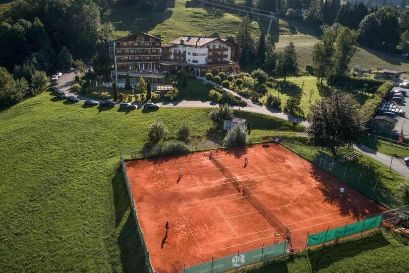 Luftaufnahme von zwei Sandtennisplätzen vor einem Hotelgebäude in Hanglage.