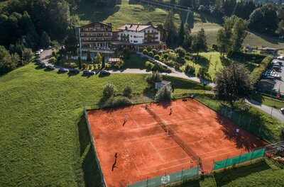 Luftaufnahme von zwei Sandtennisplätzen vor einem Hotelgebäude in Hanglage.