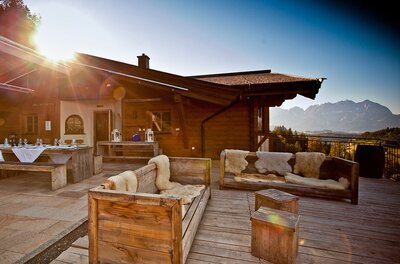 Terrasse der Ganslern Alm Holzterrasse einer Almhütte mit Sitzmöbeln aus Holz und Blick auf die Berge bei Sonnenuntergang.