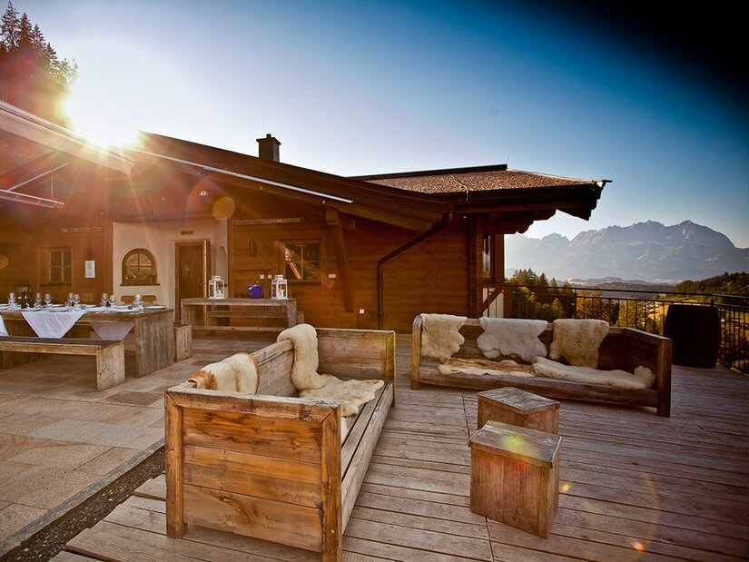 Terrasse der Ganslern Alm Holzterrasse einer Almhütte mit Sitzmöbeln aus Holz und Blick auf die Berge bei Sonnenuntergang.