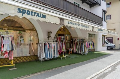 Außenansicht eines Bekleidungsgeschäfts mit Markise und Kleiderständern vor dem Eingang.