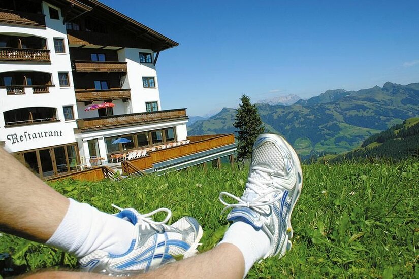 Sommer am Hotel Ehrenbachhöhe Beine in Sportschuhen auf einer Wiese vor einem Berghotel mit Blick auf die Alpen.