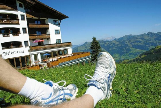 Beine in Sportschuhen auf einer Wiese vor einem Berghotel mit Blick auf die Alpen.