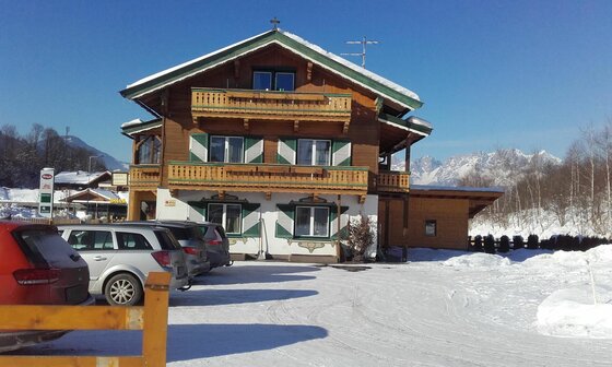 Alpines Holzhaus mit Balkonen und Fensterläden vor schneebedeckten Bergen und Parkplatz im Winter.