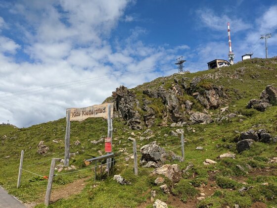 Holzportal mit der Aufschrift Kids Kletterpfad auf einer grünen Bergwiese vor felsigem Hang und Sendemast.