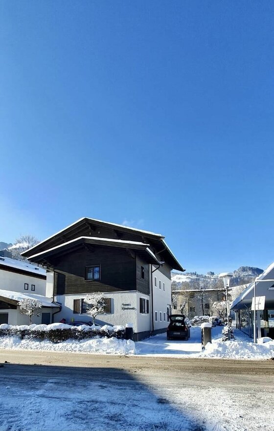 Haus Pankratz im Winter Zweistöckiges Haus mit dunkler Holzfassade im Obergeschoss in einer verschneiten Winterlandschaft unter blauem Himmel.