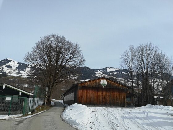 Holzscheune an einer asphaltierten Straße vor schneebedeckten Bergen und kahlen Bäumen im Winter.
