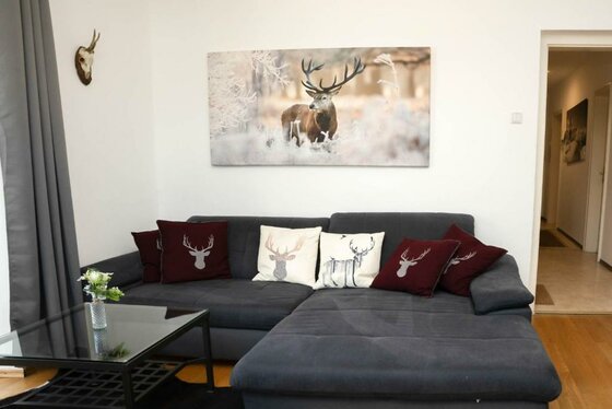 587402273 Ein gemütliches Wohnzimmer mit einer grauen Couch und dekorativen Kissen. An der Wand hängt ein Bild von einem Hirsch in naturnaher Umgebung.