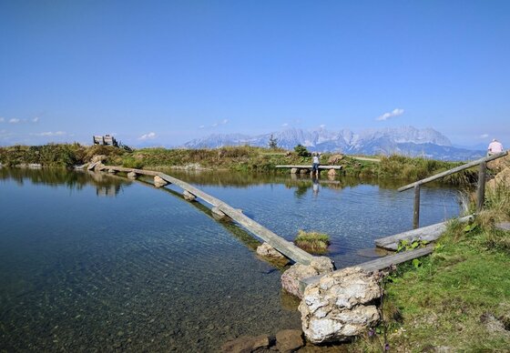 Ein schmaler Holzsteg führt über einen klaren Bergsee vor einer alpinen Gebirgskette unter blauem Himmel.