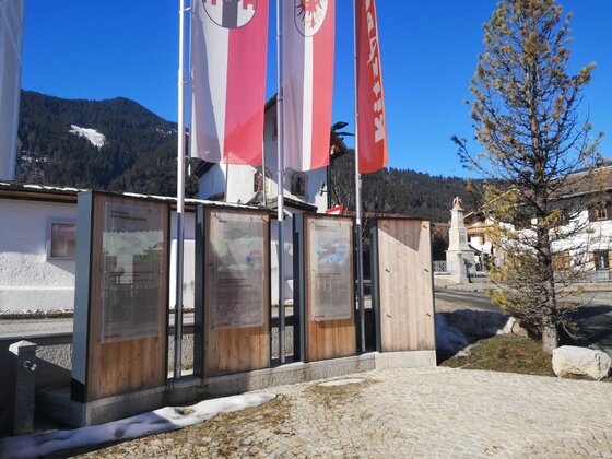 Infotafeln am Wanderweg Hennleiten Informationstafeln aus Holz und Glas vor rot-weißen Flaggen und bewaldeten Bergen.