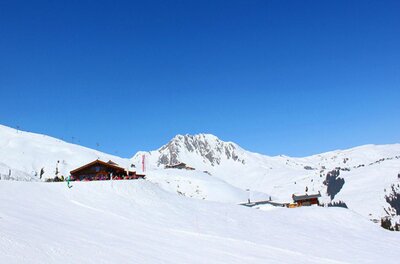 Verschneite Berghütte Hanglalm vor einem felsigen Berggipfel unter blauem Himmel.
