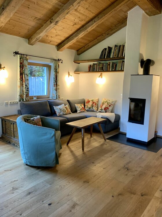 Wohnbereich im Zuhaus Schernfeichten Wohnzimmer mit grauem Ecksofa, Holztisch, Kaminofen und Bücherregalen unter einer Holzbalkendecke.