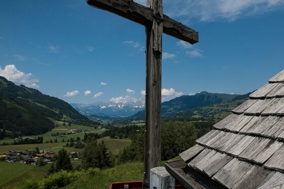 Blick auf Kochau-Hechenmoos Holzkreuz im Vordergrund vor einem grünen Tal mit Bergen unter blauem Himmel.