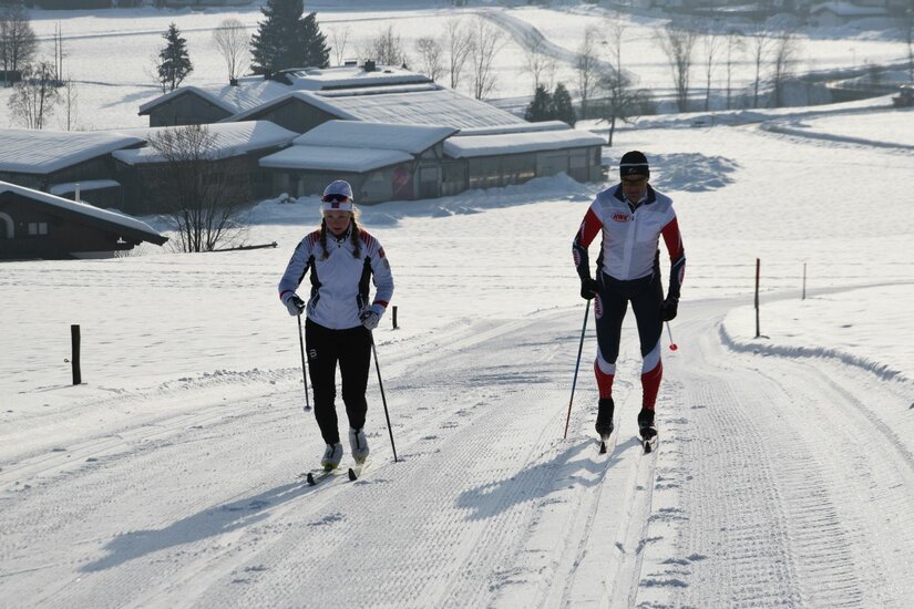 Zwei Personen beim Skilanglauf auf einer präparierten Loipe in einer verschneiten Landschaft.