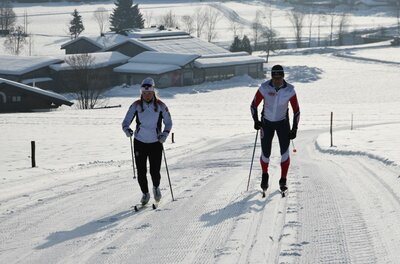 Zwei Personen beim Skilanglauf auf einer präparierten Loipe in einer verschneiten Landschaft.