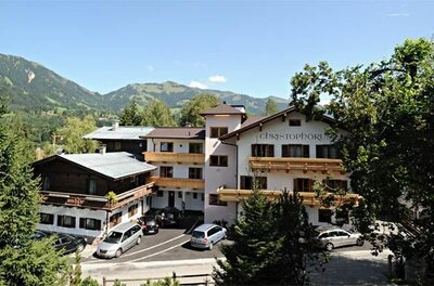 Mehrstöckiges Hotelgebäude mit Holzbalkonen und Parkplatz vor bewaldeter Bergkulisse.