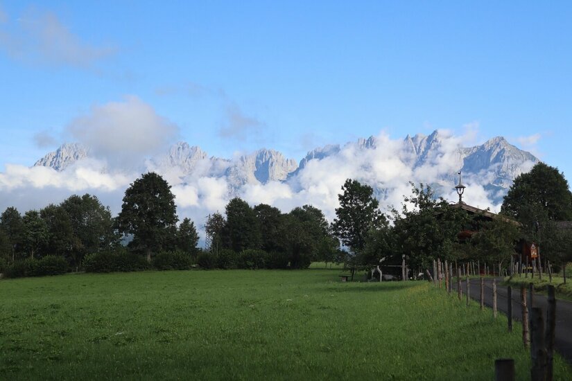 Grüne Wiese mit Bäumen vor einem wolkenverhangenen Gebirgsmassiv unter blauem Himmel.