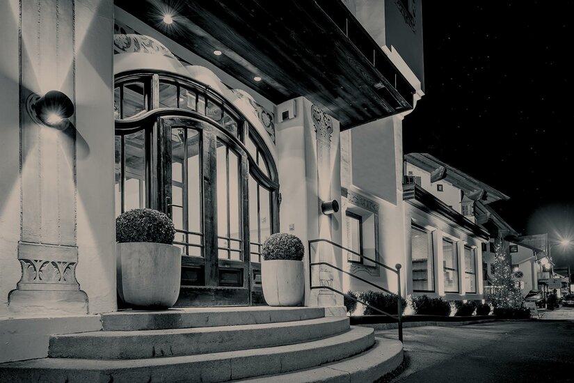 FIRST Kitzbühel Immobilien Ein eleganter Eingangsbereich eines Gebäudes bei Nacht. Die Beleuchtung und die Pflanzen schaffen eine einladende Atmosphäre.
