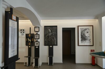 Stuck Galerie Ein moderner Ausstellungsraum mit mehreren Bildern an den Wänden. Auf der linken Seite befinden sich Informationstafeln und Stühle.