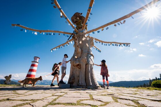Wunschbaum Eine Gruppe von Menschen steht um eine große Statue mit vielen Armen. Im Hintergrund sind Berge und ein Leuchtturm zu sehen, und die Sonne scheint.
