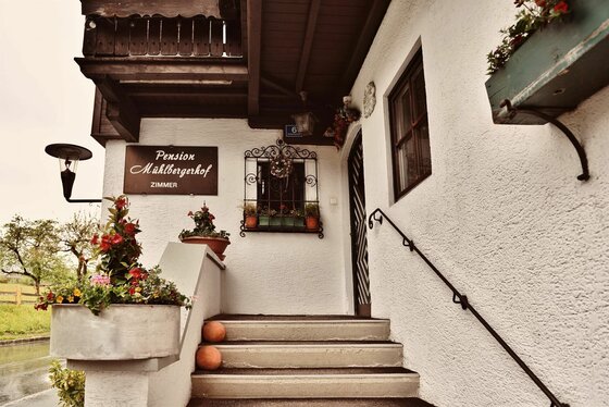 Eingang Pension Mühlbergerhof Eingangsbereich der Pension Mühlbergerhof mit Steintreppe, Blumenkästen und Namensschild an der weißen Fassade.