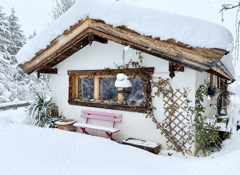 Zuhaus Schernfeichten im Winter Verschneite Hütte mit rosa Holzbank und schneebedecktem Dach bei leichtem Schneefall.