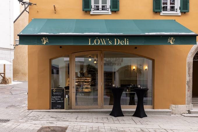 Löw's Deli Einladendes Deli mit einer grünen Markise und großen Fenstern. Der Eingang ist von einer orangefarbenen Wand umgeben, und zwei Tische stehen vor der Tür.