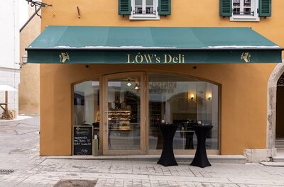 Einladendes Deli mit einer grünen Markise und großen Fenstern. Der Eingang ist von einer orangefarbenen Wand umgeben, und zwei Tische stehen vor der Tür.