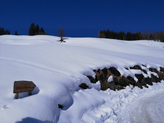 Verschneiter Hang mit Holzwegweiser zur Hagstättalm unter blauem Himmel.