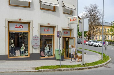 Außenansicht eines Kindermodegeschäfts mit Schaufenstern und einer Passantin mit Hund auf dem Gehweg.