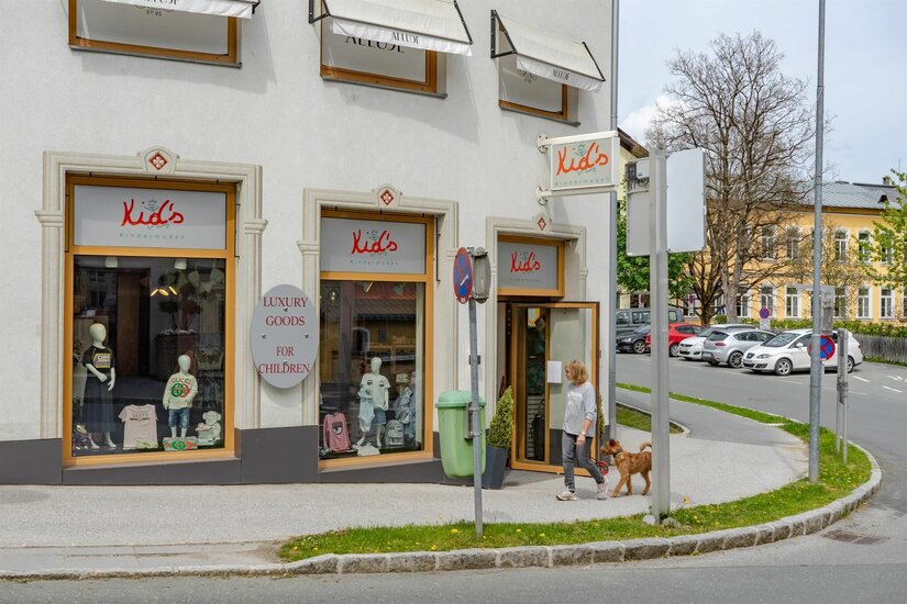 Außenansicht eines Kindermodegeschäfts mit Schaufenstern und einer Passantin mit Hund auf dem Gehweg.