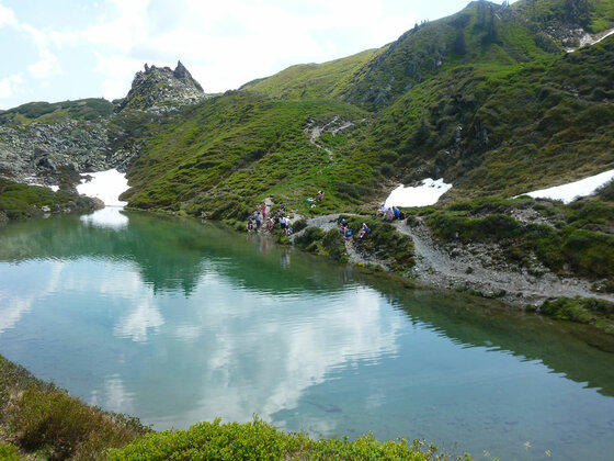 Ein klarer Bergsee umgeben von grünen Hängen, Schneeresten und Wanderern am Ufer.