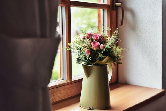 Blumenstrauß mit rosa Rosen in einer grünen Emaille-Kanne auf einem hölzernen Fensterbrett.