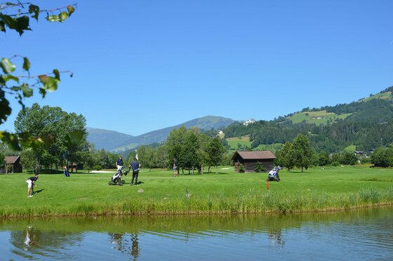 Golfplatz mit Personen am Green vor einem See und bewaldeten Bergen unter blauem Himmel.