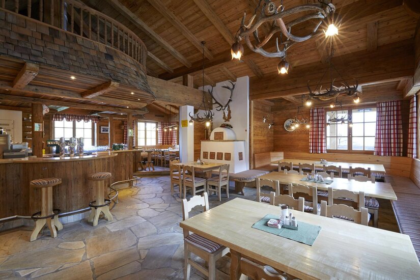 Rustikaler Gastraum mit Holztischen, Bar, Kachelofen und Geweih-Lampen.