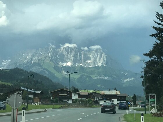 Ortsansicht mit Gebirgspanorama Blick von einer Straße auf eine Ortschaft vor einem massiven, felsigen Gebirgszug unter bewölktem Himmel.