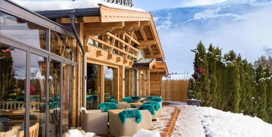 Außenterrasse einer Holzhütte mit Sesseln und blauen Fellen vor verschneiter Bergkulisse.