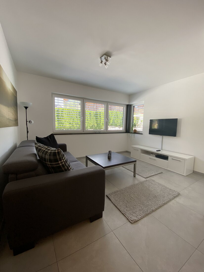 Wohnbereich im Appartement Modernes Wohnzimmer mit dunklem Sofa, Couchtisch, TV an der Wand und hellem Fliesenboden.