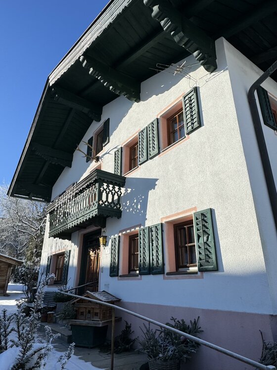Ein traditionelles Haus im alpinen Stil mit grünem Balkon und Fensterläden. Der Schnee auf dem Boden und der klare Himmel verleihen der Szene eine gemütliche Atmosphäre.