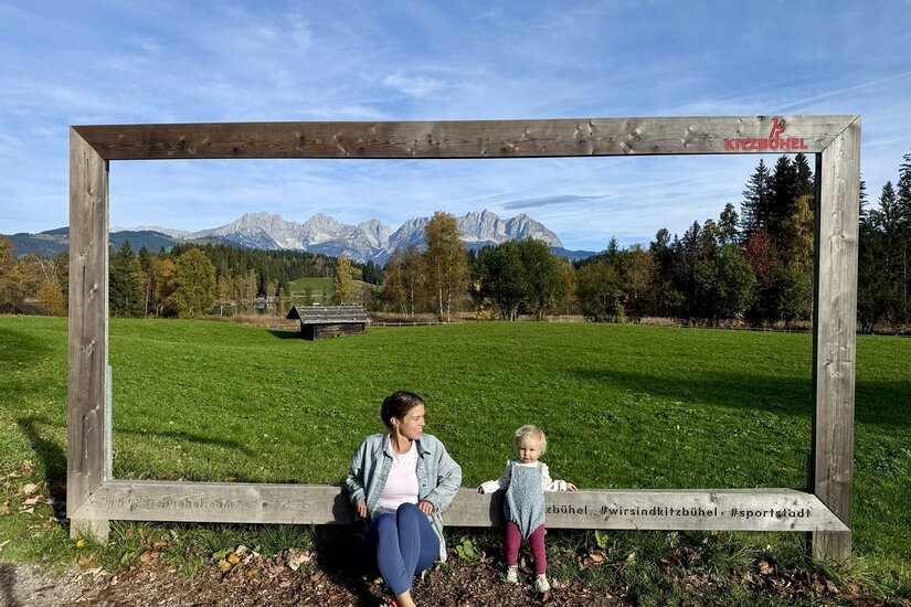Frau und Kleinkind sitzen in einem großen Holzrahmen vor einer Bergkulisse mit Wiesen und Wald.