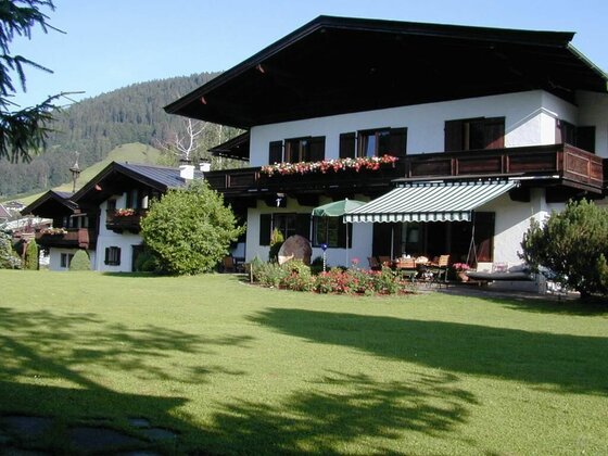 Alpines Landhaus mit großem Rasen, Blumenbalkonen und einer Terrasse mit Markise vor bewaldetem Berg.