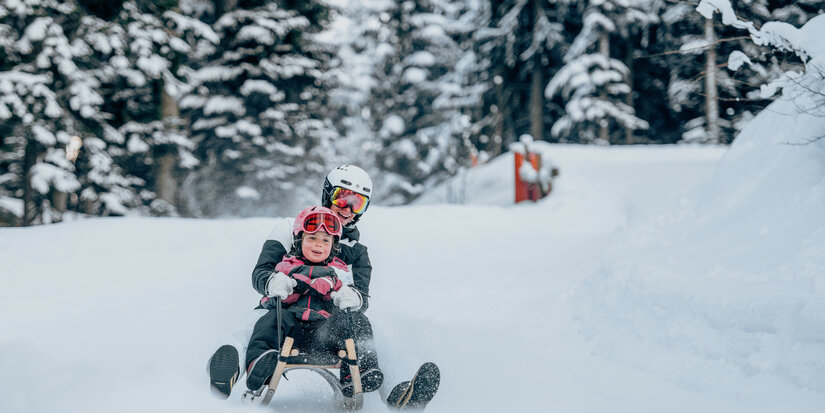 Ein Erwachsener und ein Kind fahren fröhlich auf einem Schlitten durch den verschneiten Wald, umgeben von schneebedeckten Bäumen.