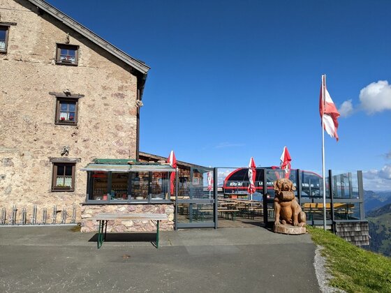 Berghütte am Hornköpflsee Berghütte aus Stein mit Terrasse, Sonnenschirmen, einer Holzskulptur und einer Seilbahnstation im Hintergrund.