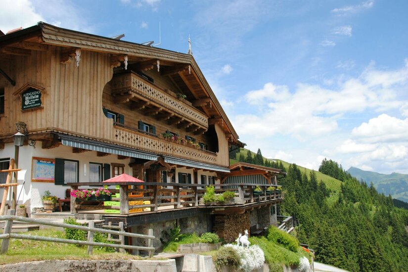 Melkalm im Sommer Alpengasthof Melkalm aus Holz mit Terrasse vor bewaldeten Berghängen unter blauem Himmel.