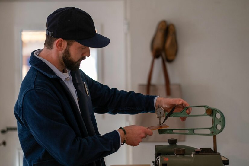 Lederhandwerk im Atelier Ein Mann mit Bart und blauer Kappe misst ein Stück Leder mit einem mechanischen Dickenmesser in einer Werkstatt.