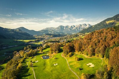 Golfclub Eichenheim Luftaufnahme eines Golfplatzes mit herbstlichen Bäumen vor einem Bergmassiv.
